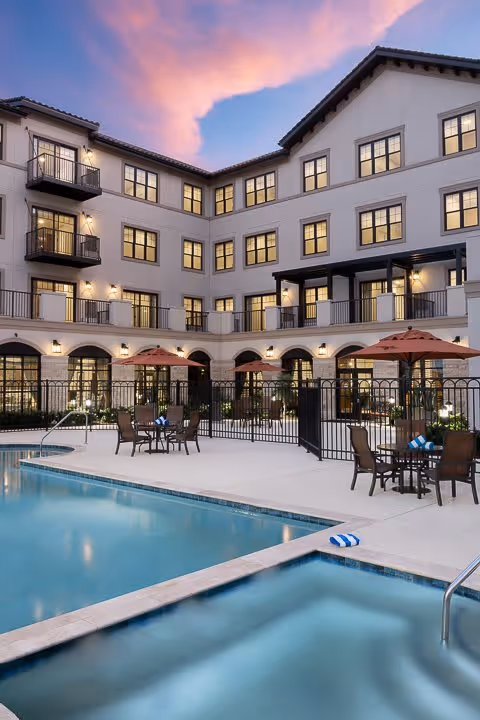 Outdoor view of a senior living facility at dusk featuring a swimming pool and hot tub in the foreground, with patio tables, chairs, and umbrellas on the pool deck. The multi-story building with balconies and lit windows surrounds the pool area under a colorful sky.