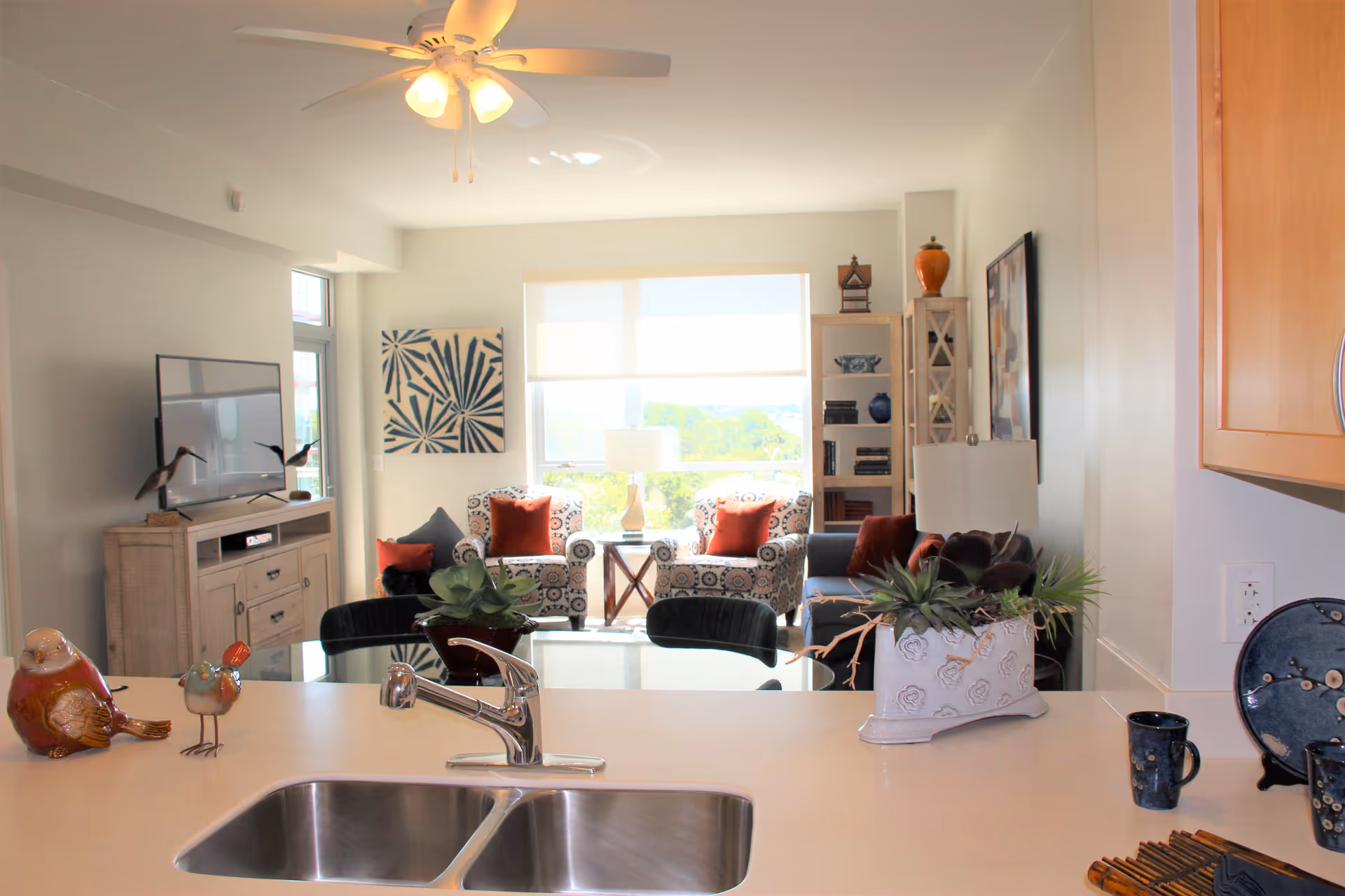 View from a kitchen counter with a double sink looking into a bright living room. The living room has two patterned armchairs with red cushions, a small table with a lamp between them, a blue sofa with red cushions, a TV on a wooden stand, and a tall bookshelf. Decorative items including plants and bird figurines are placed on the kitchen counter and TV stand.