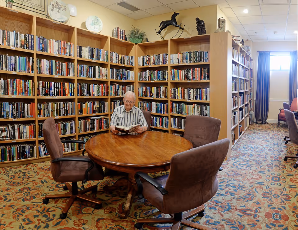 An elderly man sitting at a round wooden table reading a book in a library room with tall wooden bookshelves filled with books. The room has a patterned carpet, several brown cushioned chairs on wheels around the table, and decorative items on top of the bookshelves. A window with curtains is visible in the background.