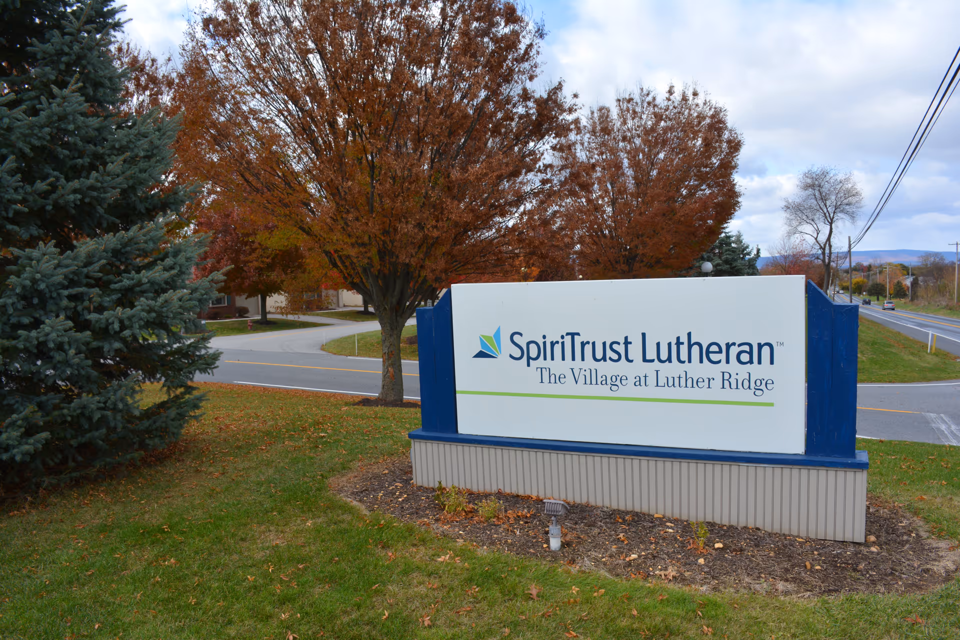 Entrance sign reading "SpiriTrust Lutheran The Village at Luther Ridge" on a grassy lawn with autumn trees and a road in the background.