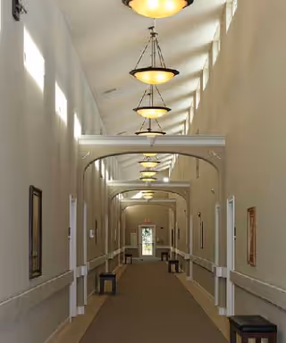 A long, well-lit hallway with beige walls and carpeted floor in a senior living community. The ceiling features multiple hanging light fixtures and small windows near the top letting in natural light. Several benches are placed along the walls, and framed pictures hang on the walls.