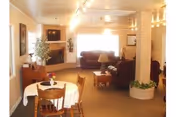 A cozy living room area with brown leather sofas, a fireplace, a small dining table with chairs, and warm lighting. There are plants and framed pictures on the walls, creating a welcoming atmosphere.