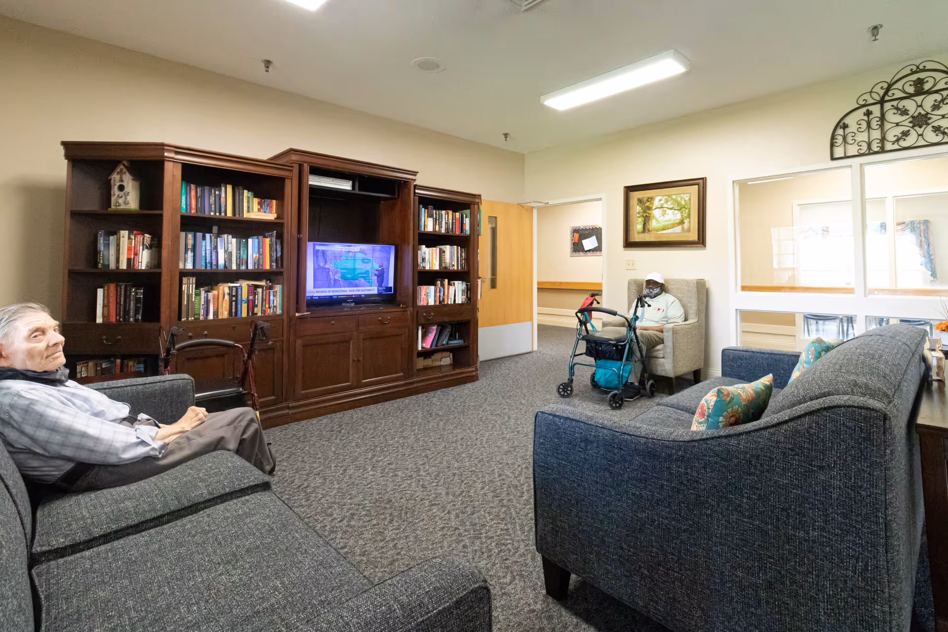 Seating area with couches facing a large bookcase and TV, with two elderly residents—one seated with a walker—relaxing in the room.