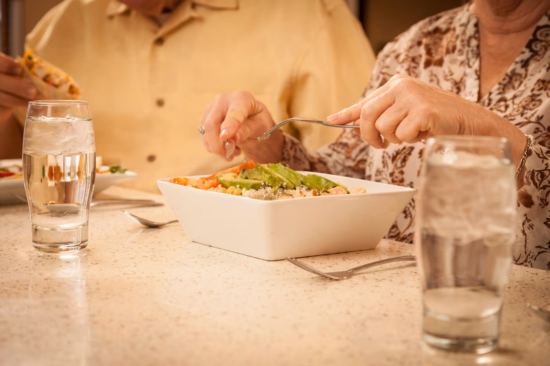 Two people eating at a table with a bowl of salad and glasses of water.