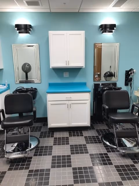 Small salon-style hair washing and styling area with two black barber chairs, sinks, mirrors, and white cabinetry against a teal wall.