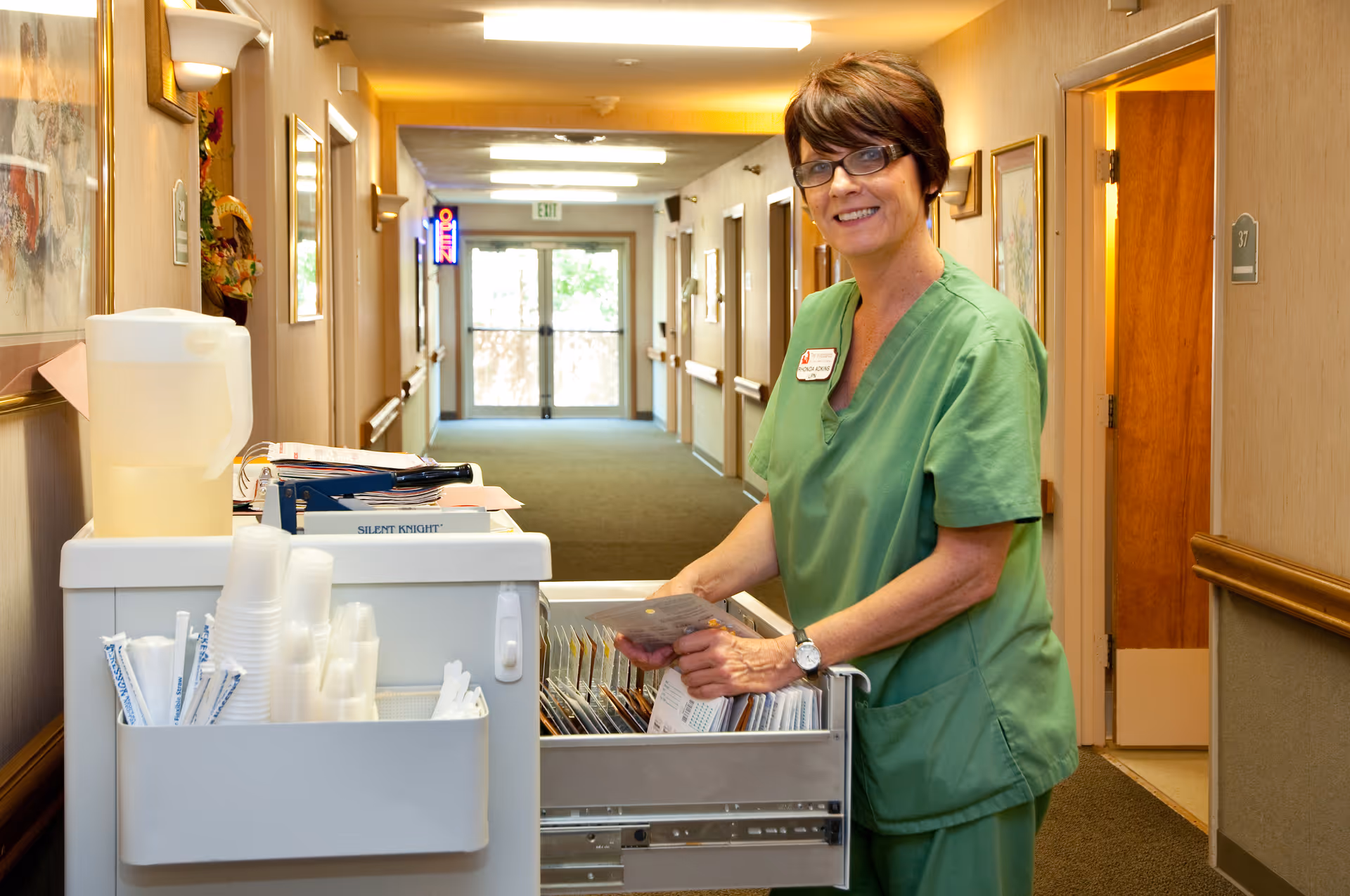 A woman in green scrubs and glasses stands in a hallway of a senior living facility, smiling while organizing items in a drawer of a mobile cart that holds cups, straws, and a pitcher of water. The hallway has beige walls, framed artwork, and several doors along the sides.