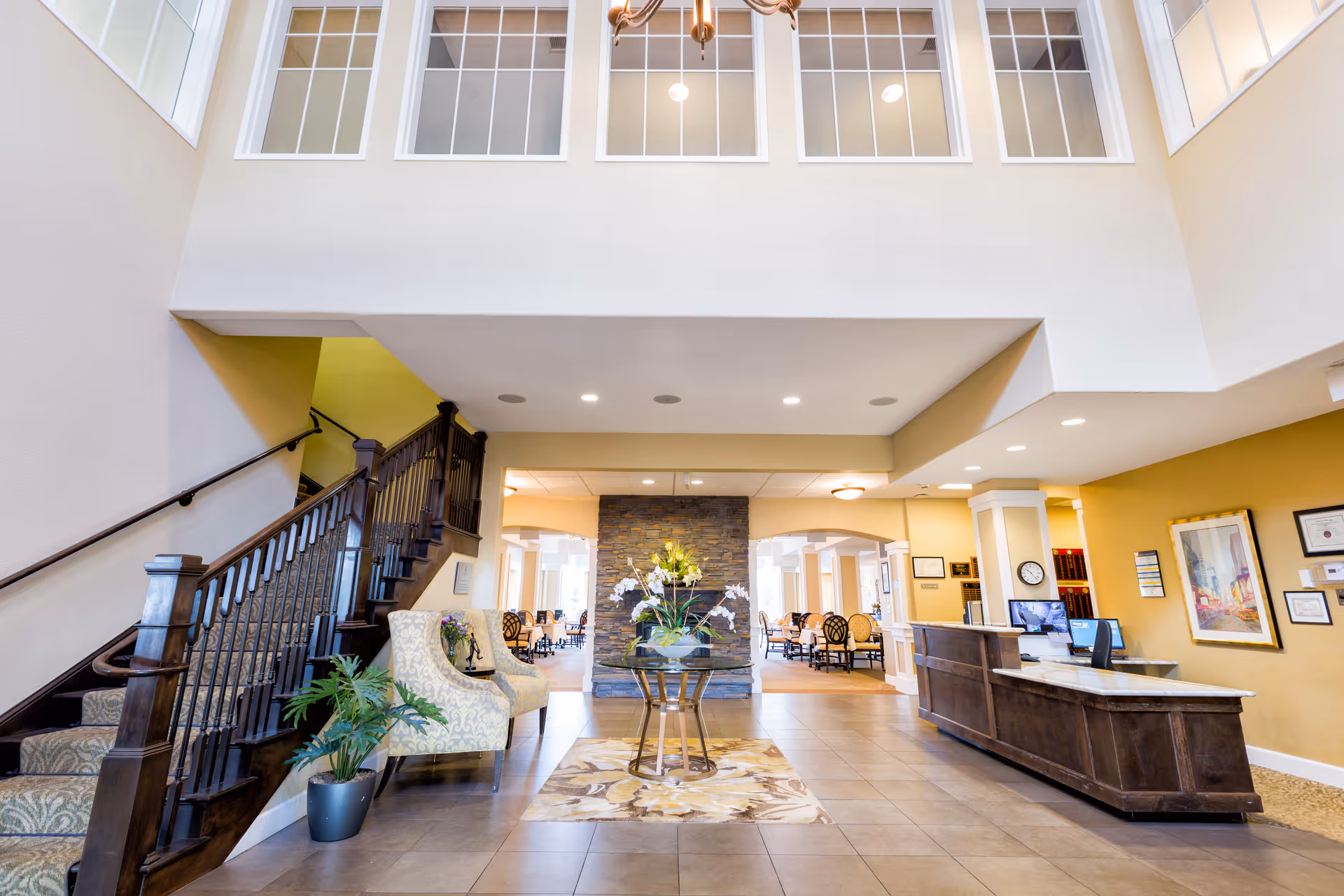 Bright and spacious assisted living facility lobby with a wooden staircase on the left, two patterned armchairs next to a potted plant, a small table with a flower arrangement in the center on a floral rug, and a reception desk on the right. The background shows a stone accent wall and a dining area with tables and chairs.