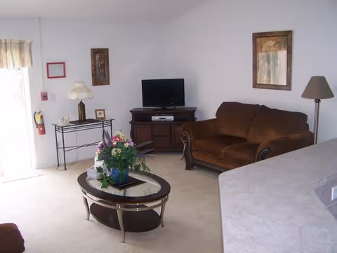 A living room with a brown couch, a glass coffee table with a floral arrangement, a TV on a wooden stand, a side table with a lamp, and framed artwork on the walls. There is a window with curtains and a fire extinguisher mounted on the wall near the door.