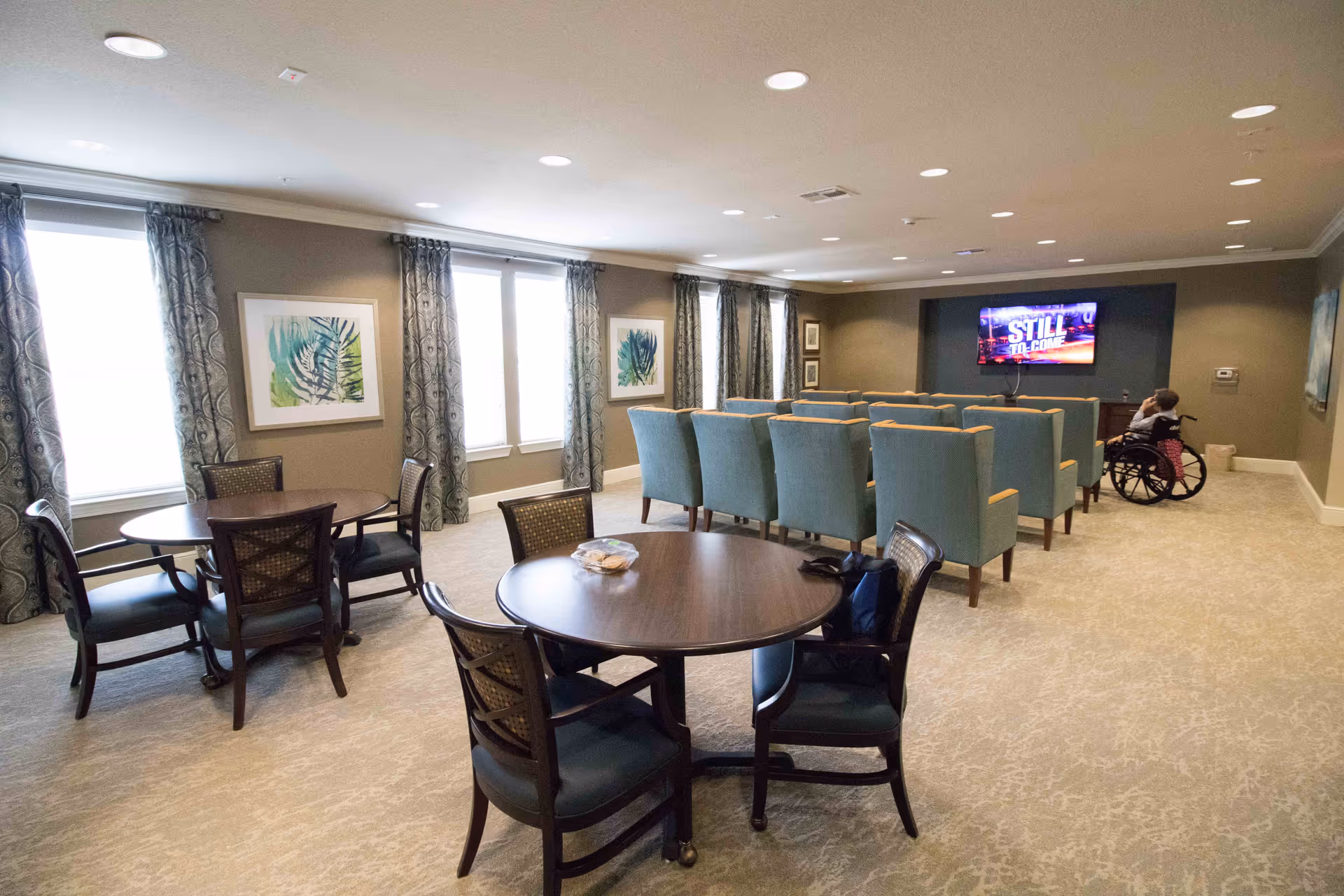 A spacious common area in an assisted living facility with round tables and chairs in the foreground and rows of blue armchairs facing a wall-mounted television in the background. A person in a wheelchair is seated near the TV. The room has large windows with patterned curtains and framed artwork on the walls.