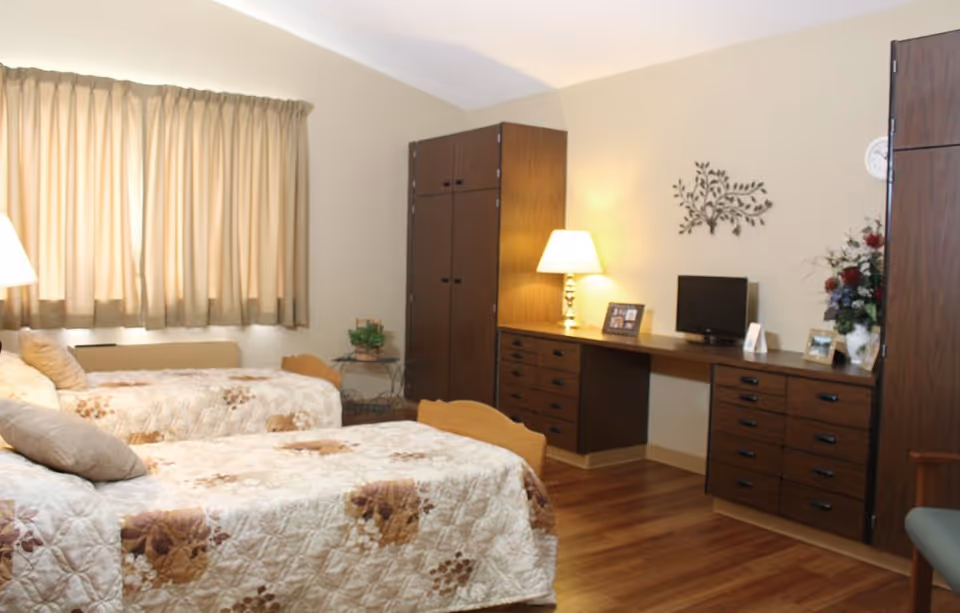A bright bedroom with two twin beds with floral quilts, wooden dressers and a desk with a lamp and small TV.