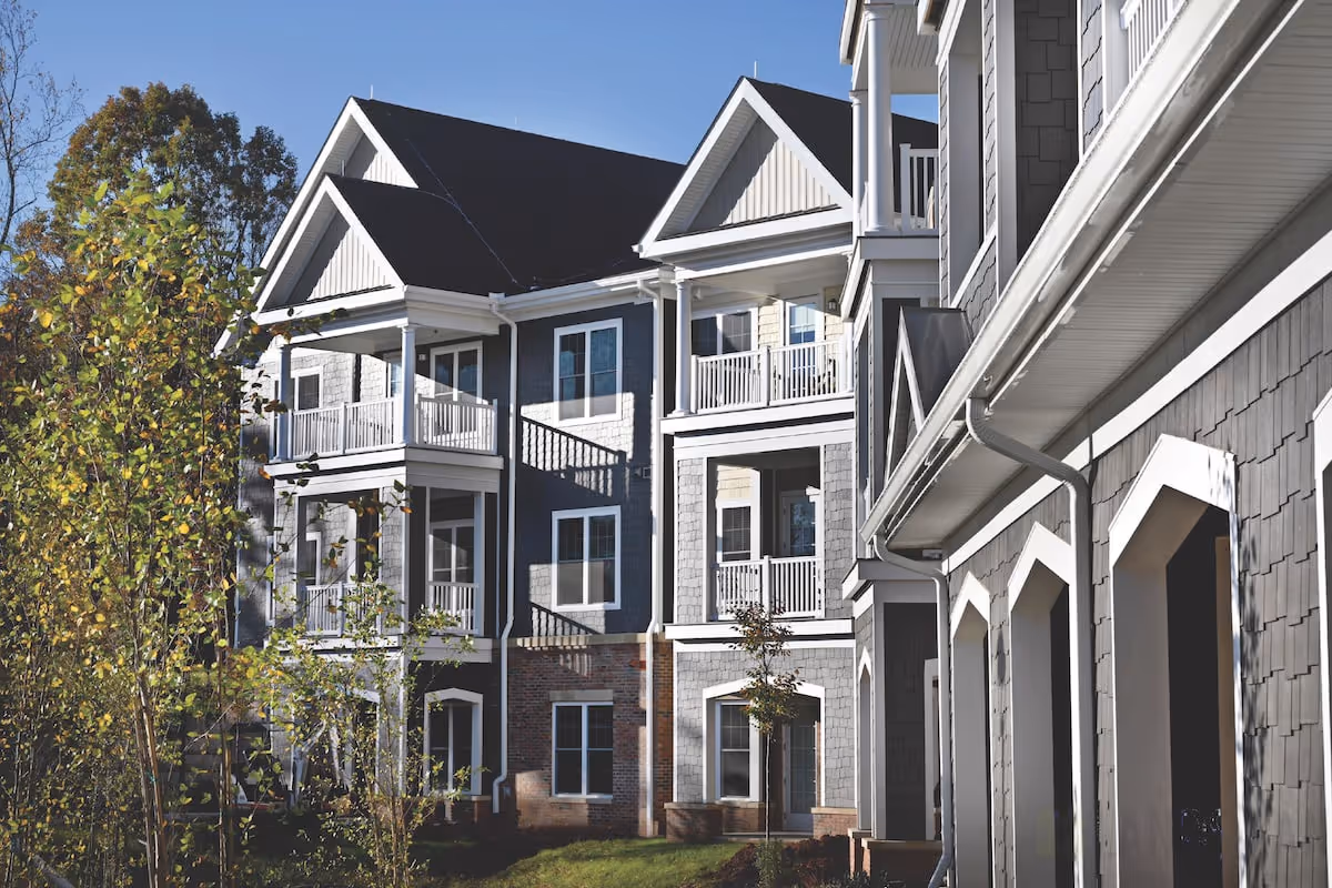 Exterior view of a multi-story senior living community building with gray siding, white trim, balconies, and a pitched roof under a clear blue sky. Trees and grass are visible in the foreground.