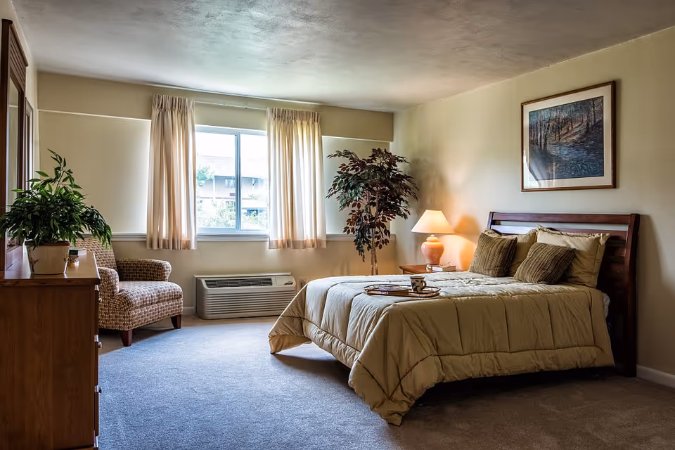 Cadbury at Cherry Hill senior living bedroom with bed, plants, chair, dresser, and window.