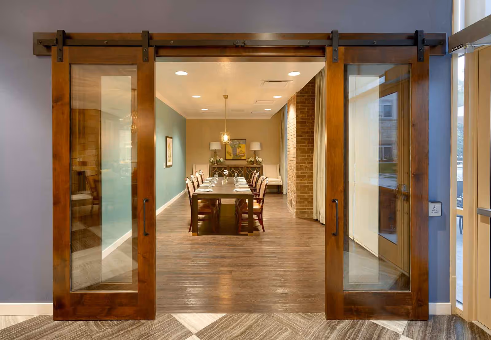 View through large wooden sliding glass doors into a dining room with a long table set for eight people. The room has wooden flooring, a blue accent wall on the left, a brick wall on the right, and two white chairs and a sideboard with lamps and artwork at the far end.