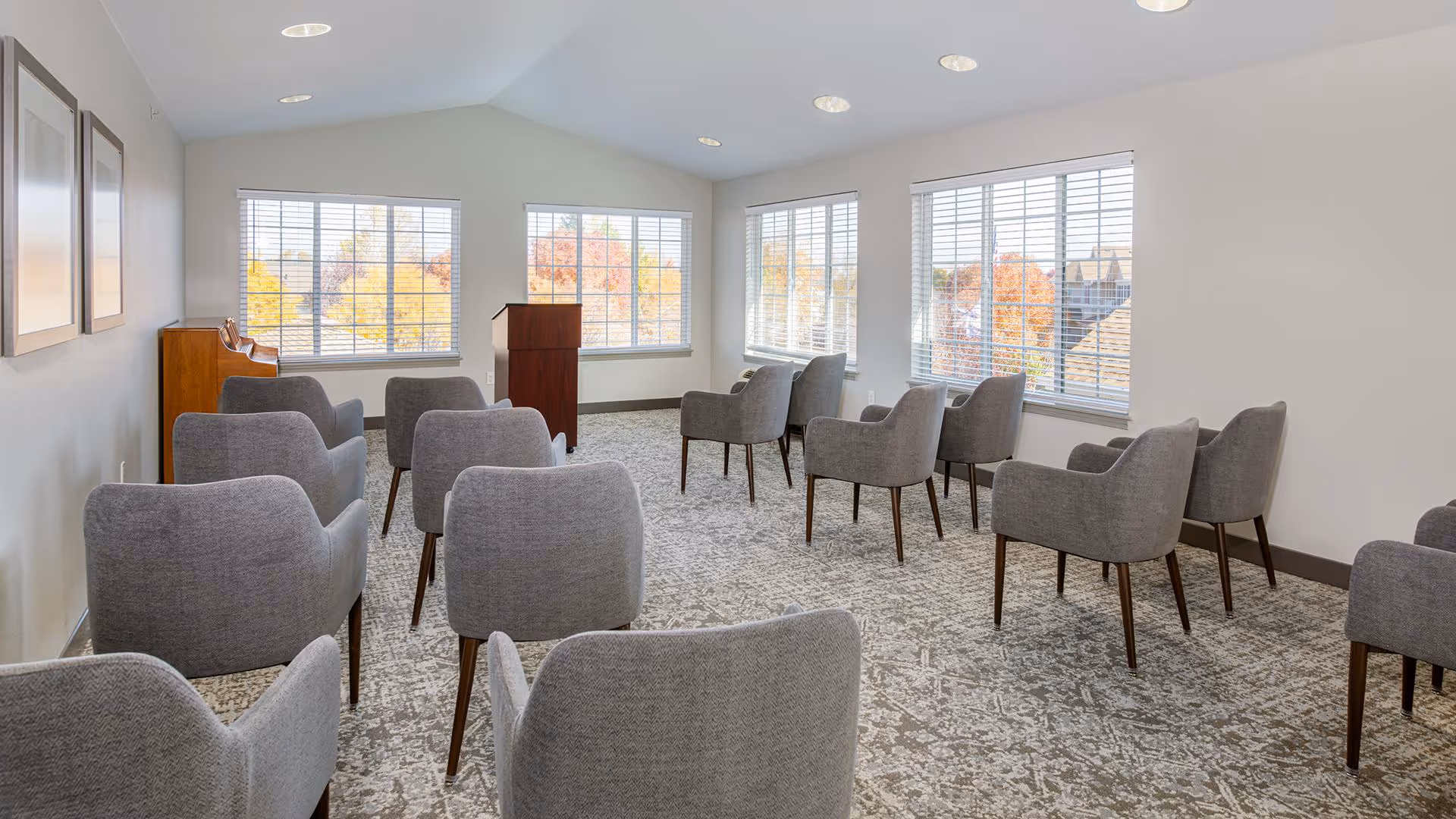 A bright, modern meeting or presentation room with multiple gray upholstered chairs arranged in rows facing a wooden podium. The room has large windows with white blinds letting in natural light and a patterned carpet floor.