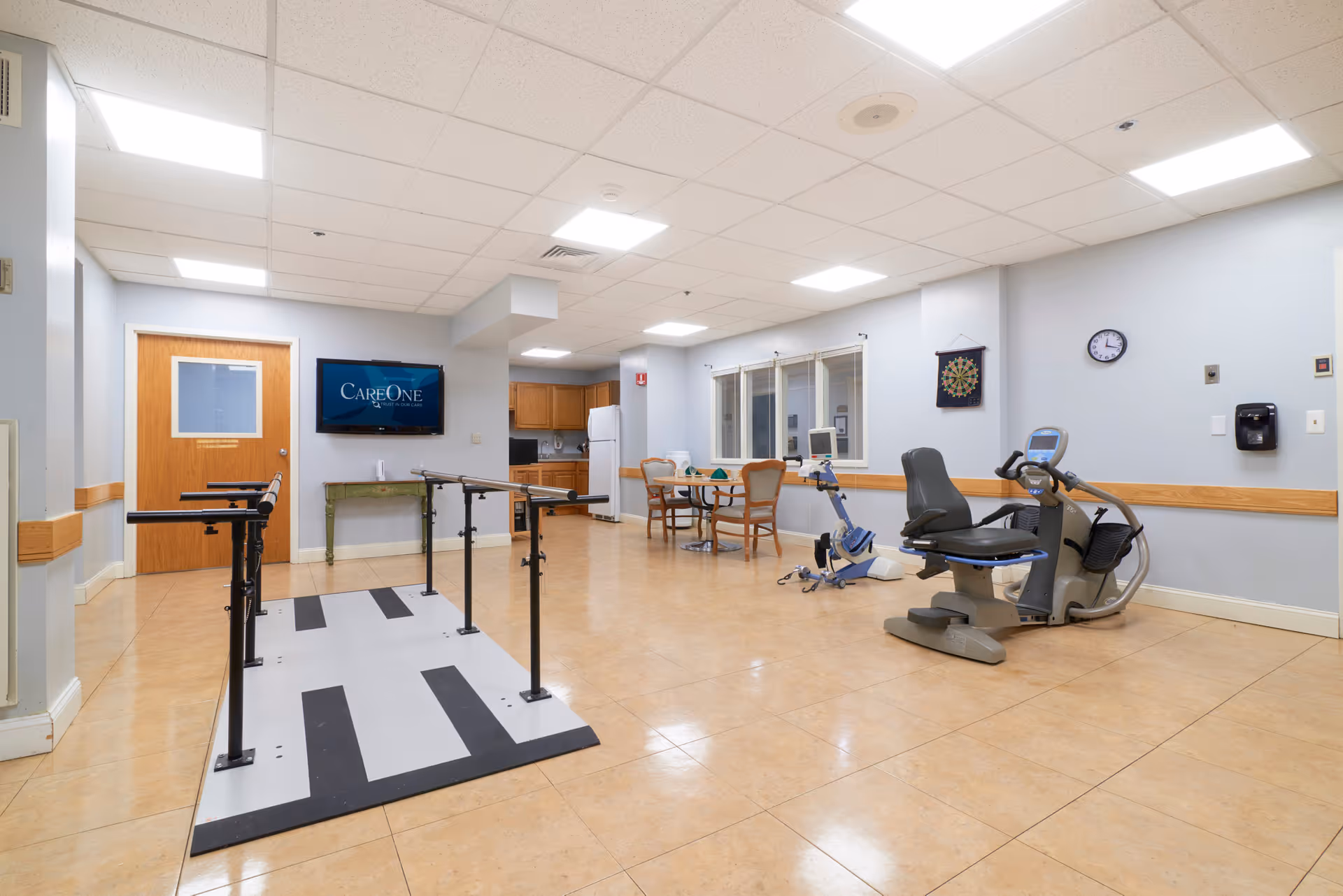 A spacious rehabilitation or therapy room in a senior living facility with parallel bars for walking exercises, a recumbent exercise bike, a small table with chairs, a dartboard on the wall, and a wall-mounted TV displaying the CareOne logo. The room has light-colored tiled floors, light blue walls with wooden chair rails, and a drop ceiling with fluorescent lighting.