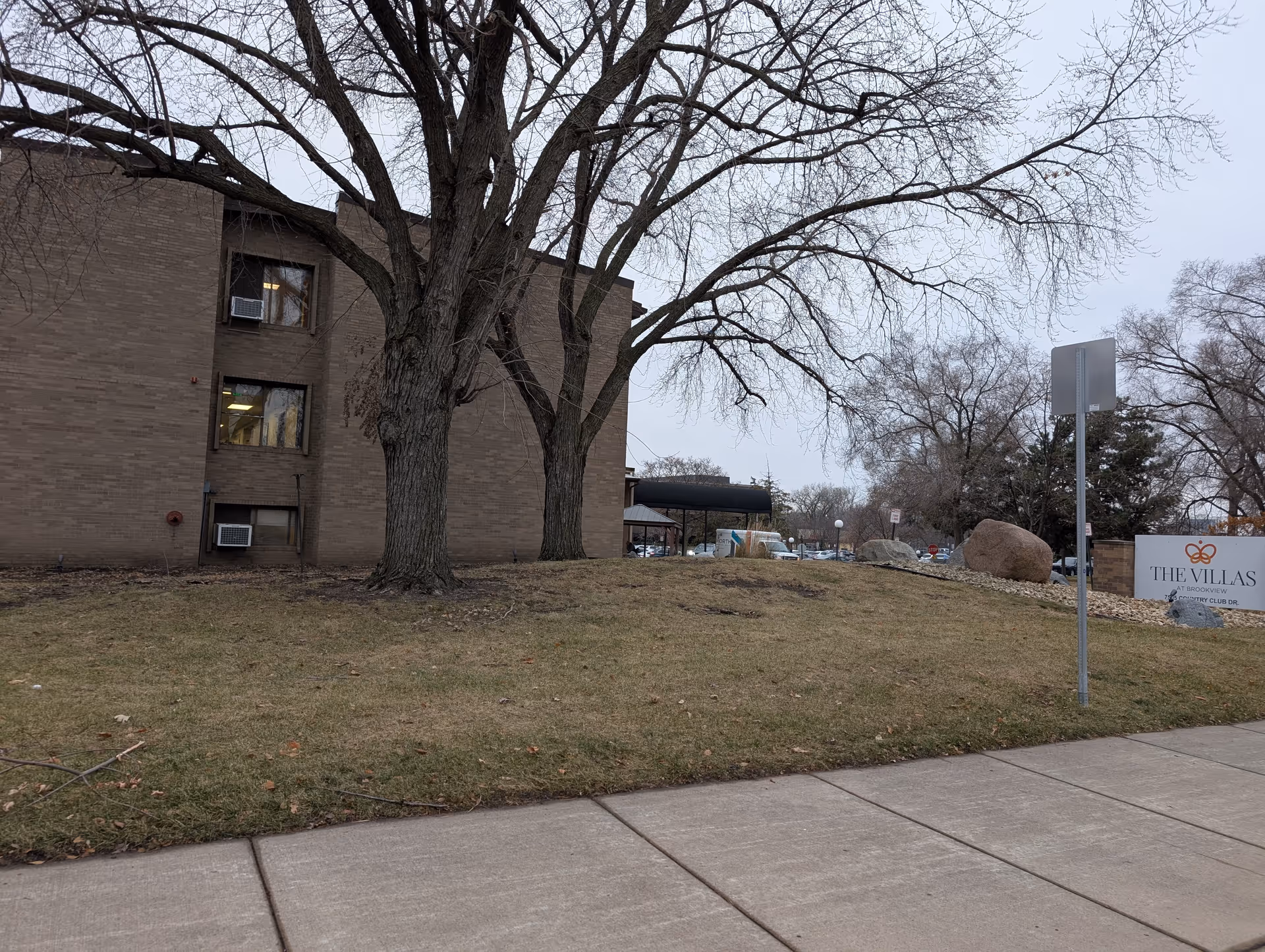 Exterior view of The Villas at Brookview building with leafless trees on a grassy area and a sidewalk in the foreground. A sign with the facility name is visible near the right side of the image.