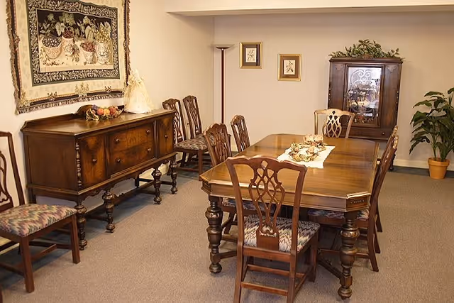 A traditional dining room with a wooden dining table surrounded by six matching chairs with patterned cushions. There is a wooden sideboard against the wall with decorative items on top, a tapestry hanging above it, and a tall floor lamp in the corner. A wooden cabinet with glass doors and a potted plant are visible in the background.
