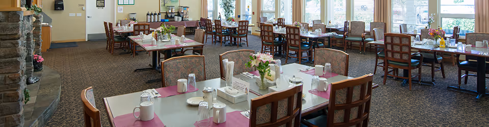 A spacious dining room with multiple tables set for meals, each table featuring placemats, cups, glasses, napkins, and flower centerpieces. The room has large windows allowing natural light to fill the space, and there is a stone fireplace on the left side.