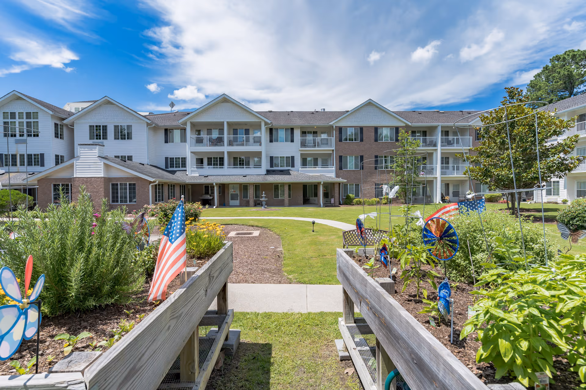 View of the exterior of a senior living facility building with three stories, balconies, and a garden area in the foreground featuring raised garden beds with plants, American flags, and decorative pinwheels under a partly cloudy blue sky.