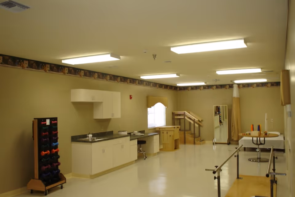 A spacious room with beige walls and ceiling lights, featuring a countertop with cabinets, a rack of colorful dumbbells, a small set of stairs with handrails, a full-length mirror, parallel bars for walking assistance, and a table with therapy equipment, suggesting a physical therapy or rehabilitation area.