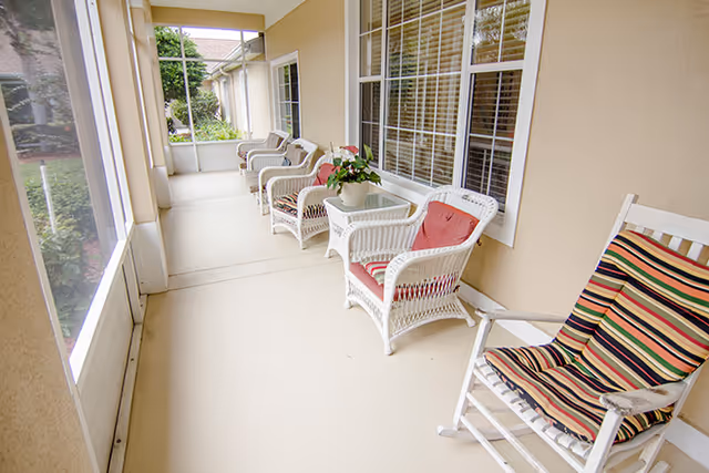 A screened-in porch with white wicker chairs and a rocking chair, some with colorful cushions, and a small table with a potted plant. The porch overlooks a garden area through the screened windows.