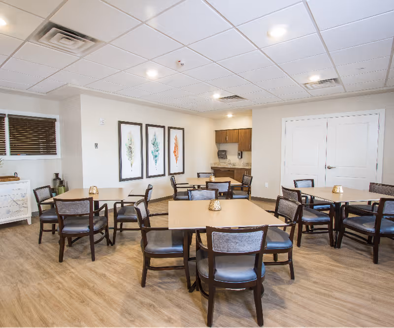 Well-lit communal dining room with several square tables and chairs, framed artwork on the wall, and a small kitchenette area in the back.