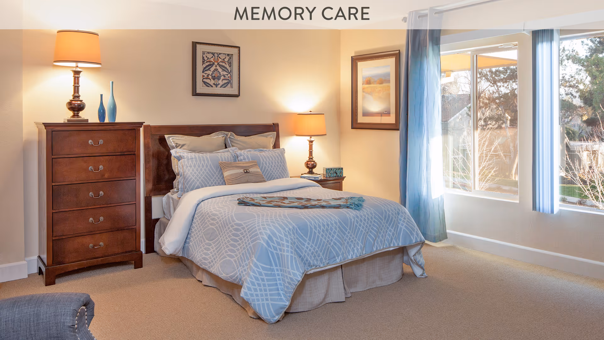 A well-lit bedroom with a neatly made bed featuring blue and white patterned bedding. There is a wooden dresser with a lamp and decorative vases on the left side of the bed, and a nightstand with a lamp and a small decorative item on the right side. The room has beige walls, carpeted floor, two framed pictures on the walls, and large windows with blue curtains letting in natural light. The word 'MEMORY CARE' is displayed at the top center of the image.