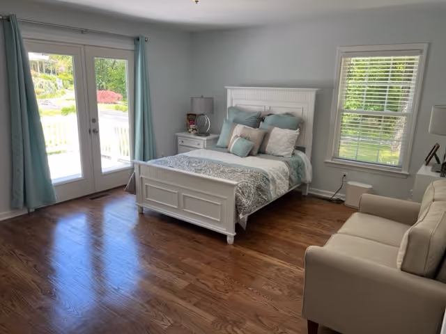 A bright bedroom with a white wooden bed frame, neatly made with blue and white bedding and multiple pillows. There is a white nightstand with a lamp and decorative items next to the bed. A beige armchair is positioned near a window with white blinds. French doors with light blue curtains open to an outdoor area with greenery visible outside. The room has wooden flooring and light-colored walls.