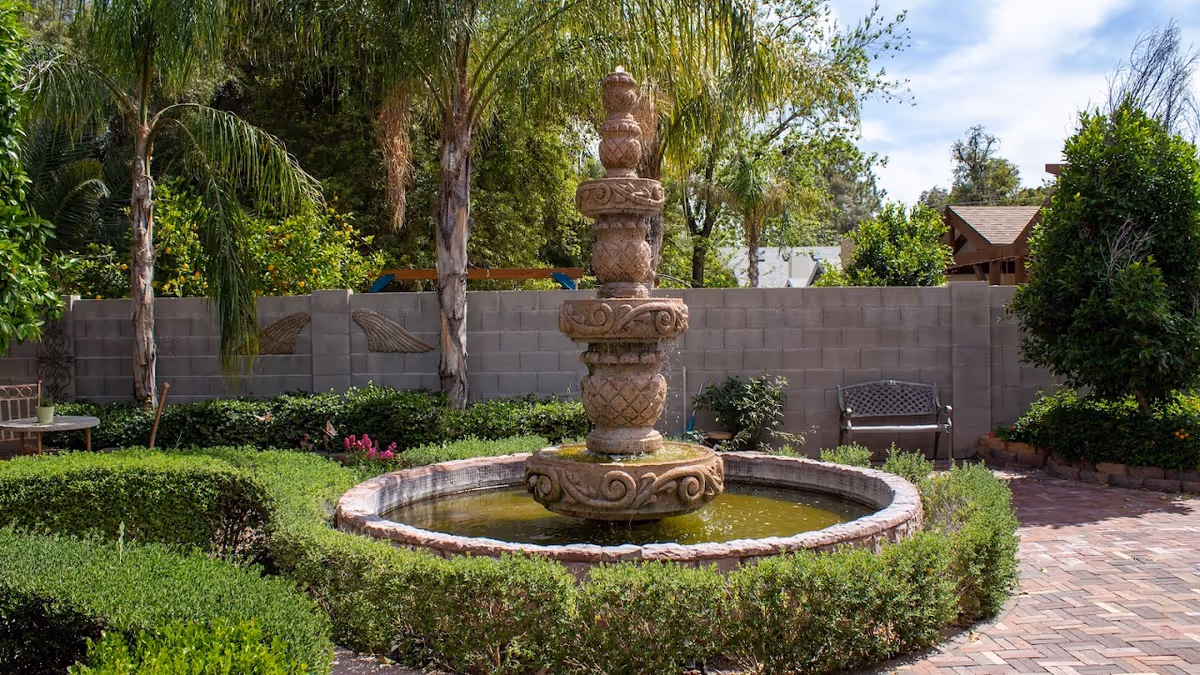 A serene outdoor garden area featuring a decorative stone fountain surrounded by neatly trimmed hedges. There are palm trees, various green plants, and a brick-paved walkway. A bench is placed against a gray block wall in the background under a partly cloudy sky.