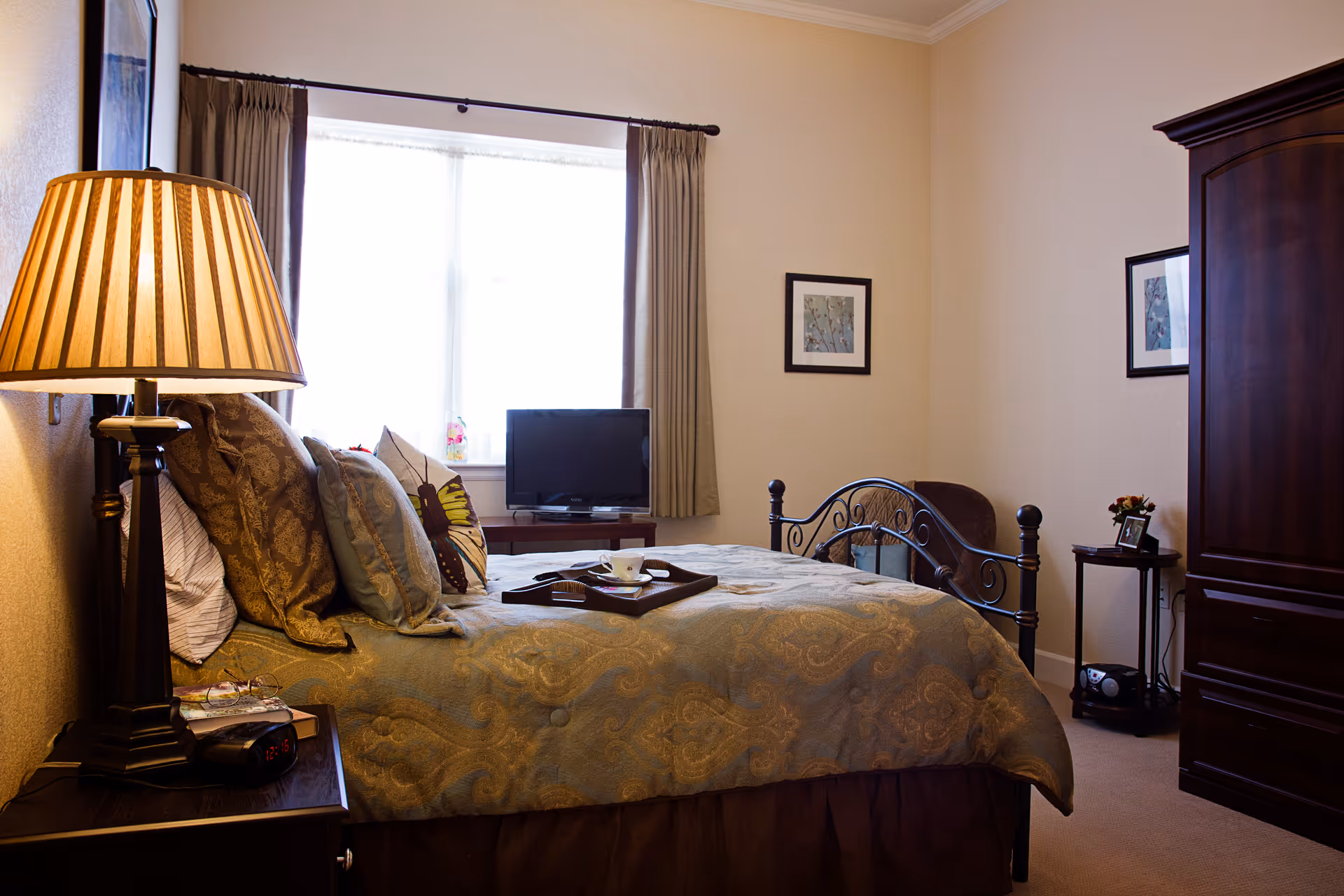 Cozy bedroom with a made bed, decorative pillows, a bedside lamp, television by the window, and a wooden armoire.