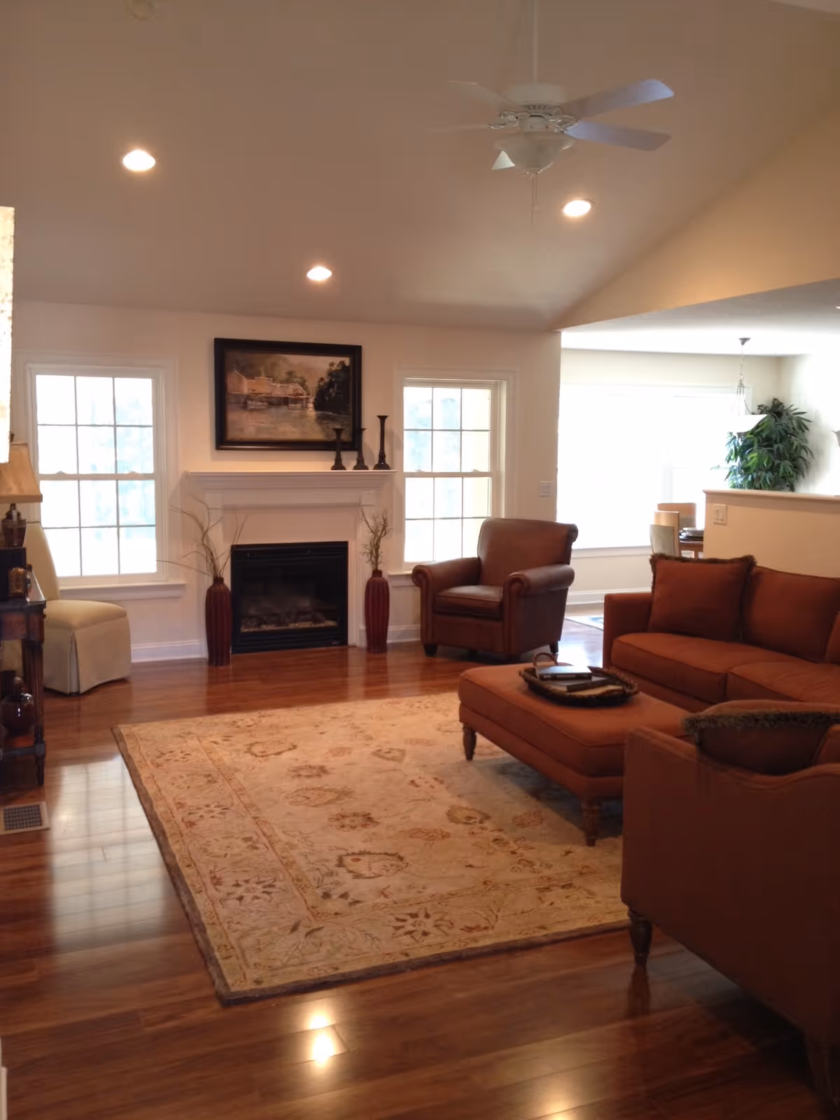 A cozy living room with wooden flooring, a large beige patterned rug, a brown leather armchair, a matching brown sofa with cushions, and an ottoman. There is a white fireplace with decorative vases and a framed painting above it, flanked by two windows. The ceiling has recessed lighting and a white ceiling fan. In the background, a dining area with a plant is partially visible.