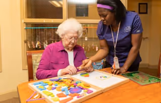 An elderly woman in a purple jacket sitting at a table engaged in a puzzle activity with a caregiver wearing a navy blue uniform who is assisting her.