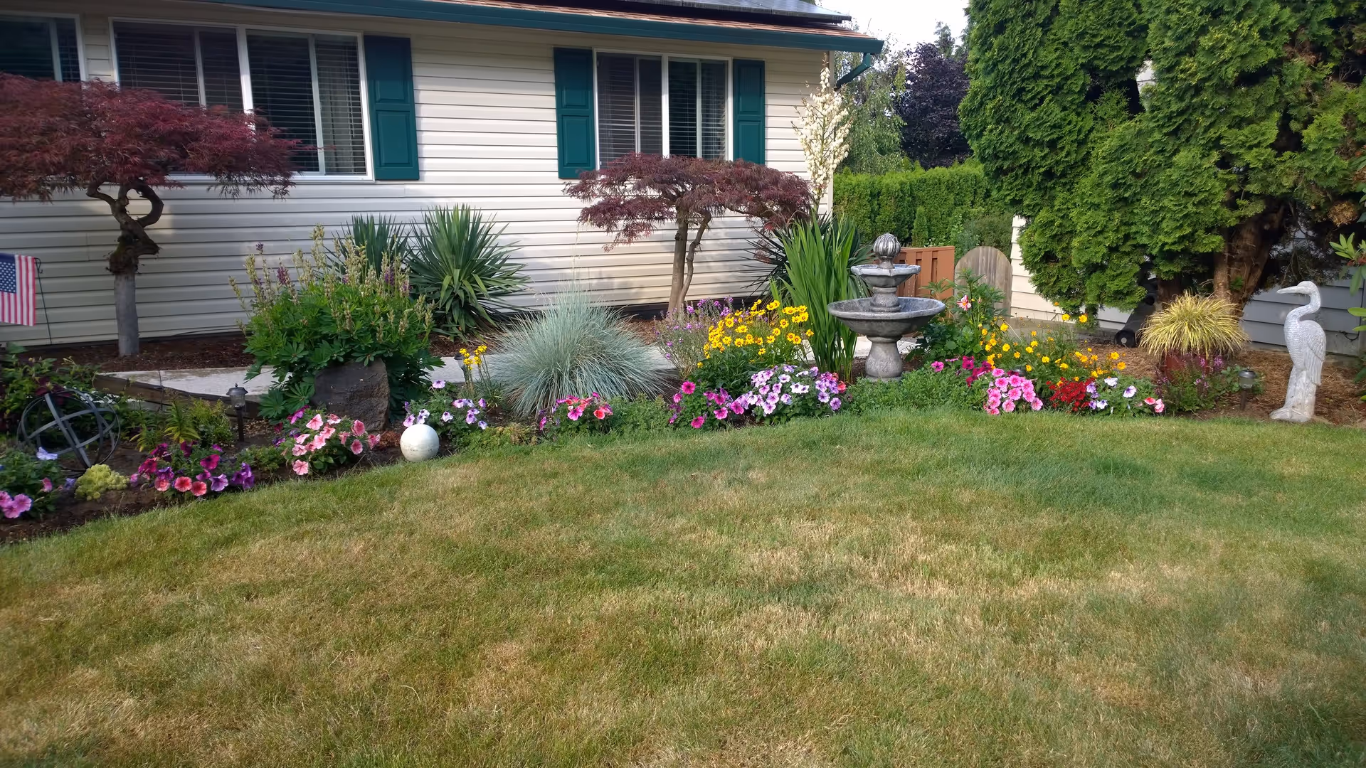 A well-maintained garden area in front of a house with beige siding and green shutters. The garden features a variety of colorful flowers, small trees, shrubs, a stone bird statue, and a tiered water fountain. The lawn in the foreground is green with some dry patches.
