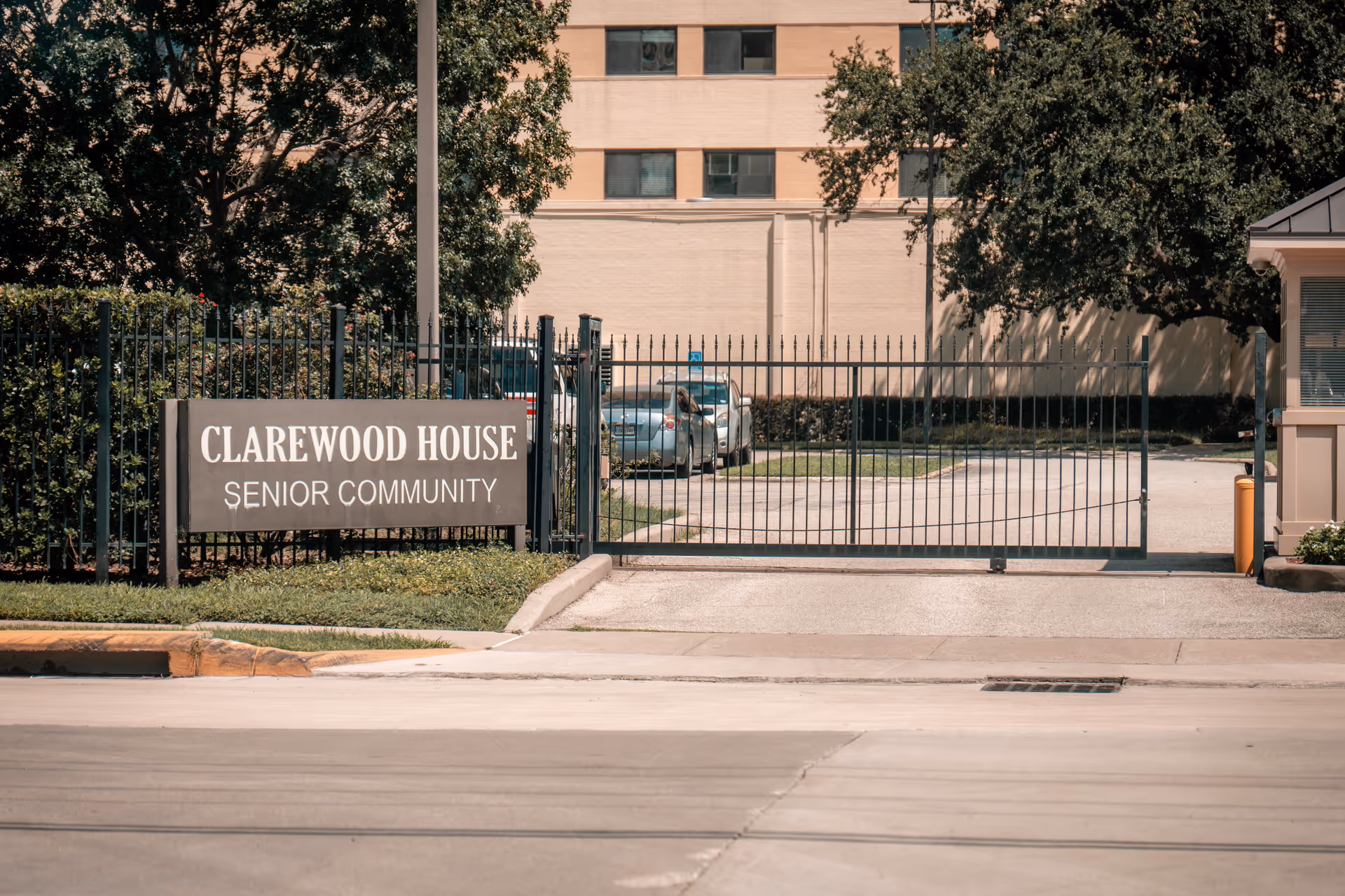 Entrance gate to Clarewood House Senior Community with a metal fence and a sign displaying the facility name. There are trees and parked cars visible inside the gated area.