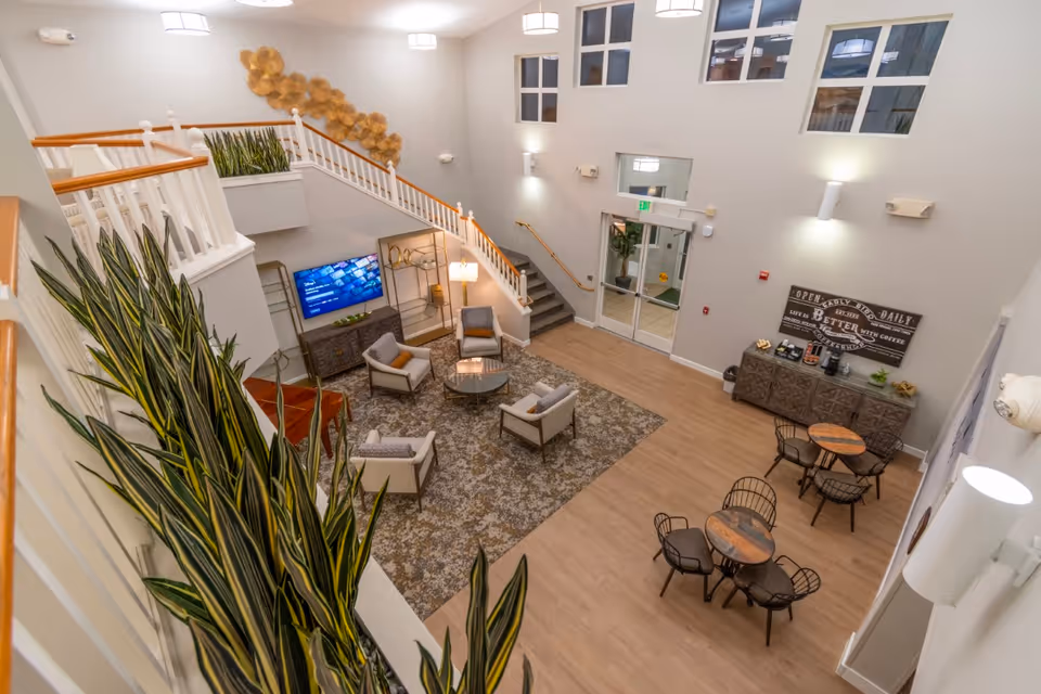 Interior view of a senior living facility common area with a seating arrangement of four armchairs around a glass coffee table on a patterned rug. There is a TV mounted on the wall, a staircase with white railings and wooden handrails, and several potted plants. To the right, there are small round tables with chairs and a sideboard with coffee and tea supplies. The space has high ceilings with multiple windows and modern light fixtures.