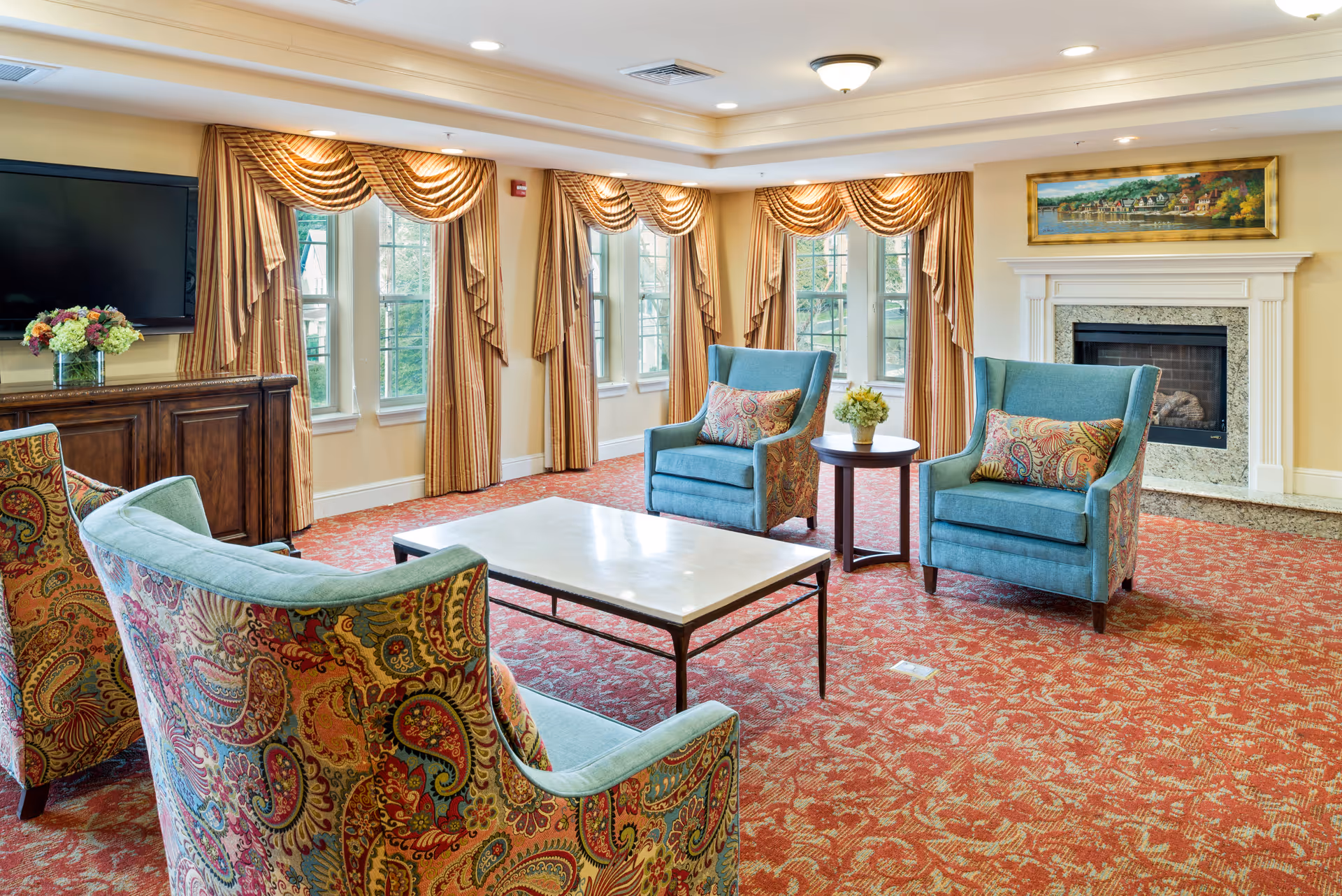 Bright lounge with patterned and blue armchairs around a coffee table, fireplace, TV, and draped windows.