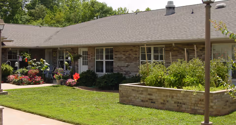 Single-story brick building with multiple windows and a door, surrounded by a well-maintained garden with various plants and flowers, and a grassy lawn in the foreground under a clear sky.
