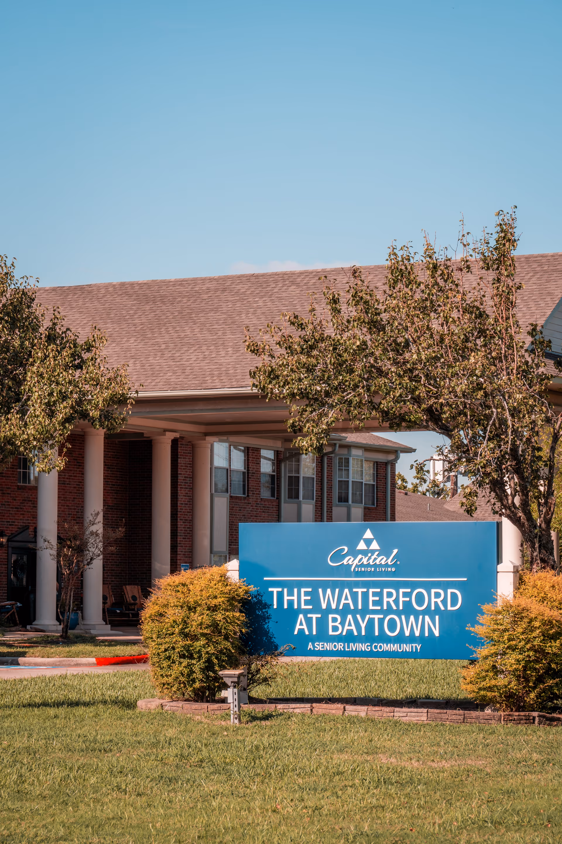 Front exterior of The Waterford at Baytown senior living facility with a blue sign on the lawn and the building entrance behind it.