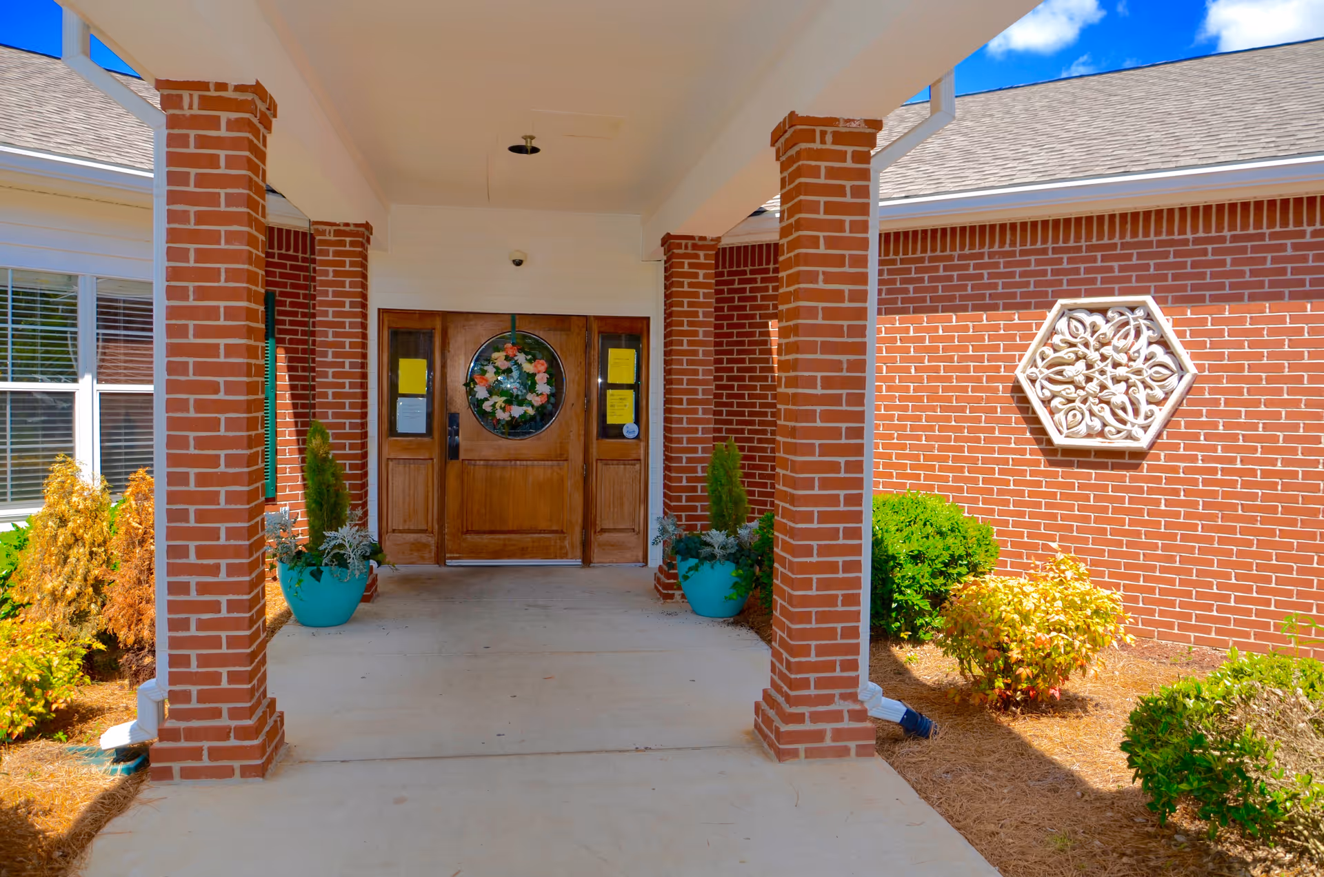 Covered entrance to a brick building with wooden double doors featuring a circular floral decoration. The entrance is flanked by brick columns and potted plants, with landscaped bushes and a decorative wall ornament on the right side under a blue sky with clouds.