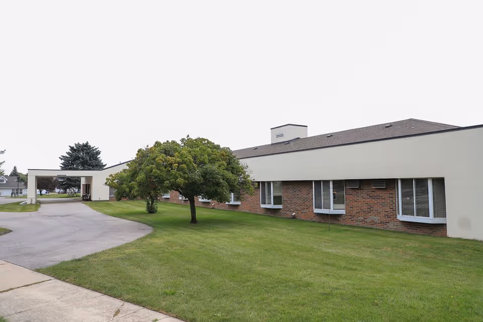 Exterior view of a single-story assisted living facility building with a brick and light-colored facade, several windows, a tree on a grassy lawn, and a driveway leading to a covered entrance.
