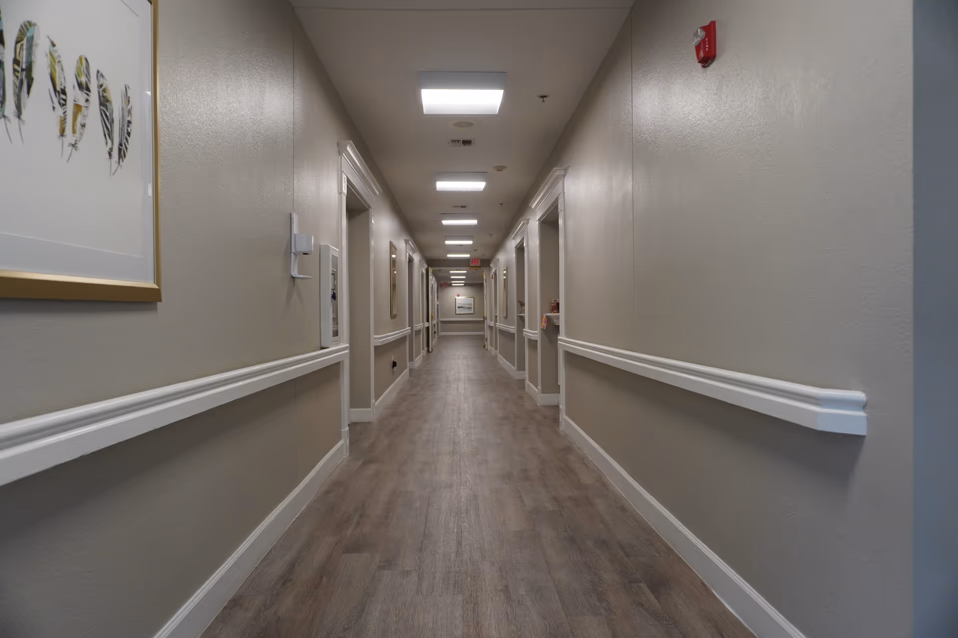 A long, well-lit hallway in a senior living facility with beige walls, white trim, and wood-look flooring. Several doorways line both sides of the corridor, and framed artwork is visible on the walls.