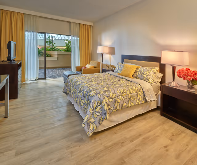 A spacious bedroom in Melrose Assisted Living featuring a large bed with floral patterned bedding and multiple pillows. The room has wooden flooring, a bedside table with a lamp and a vase of flowers, an armchair with an ottoman, a TV on a dresser, and sliding glass doors leading to an outdoor patio with plants. Yellow curtains frame the glass doors.