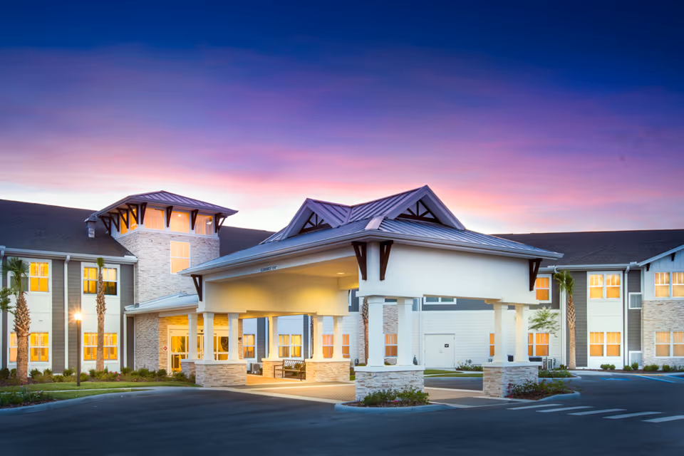 Exterior view of The Crossings at Riverview senior living facility at dusk, showing a large covered entrance with columns, well-lit windows, palm trees, and a colorful sunset sky in the background.