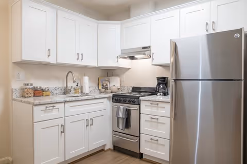 A modern kitchen with white cabinets, granite countertops, a stainless steel refrigerator, a stainless steel gas stove with an oven, a coffee maker, and various kitchen items on the counter including jars of food and a paper towel holder.