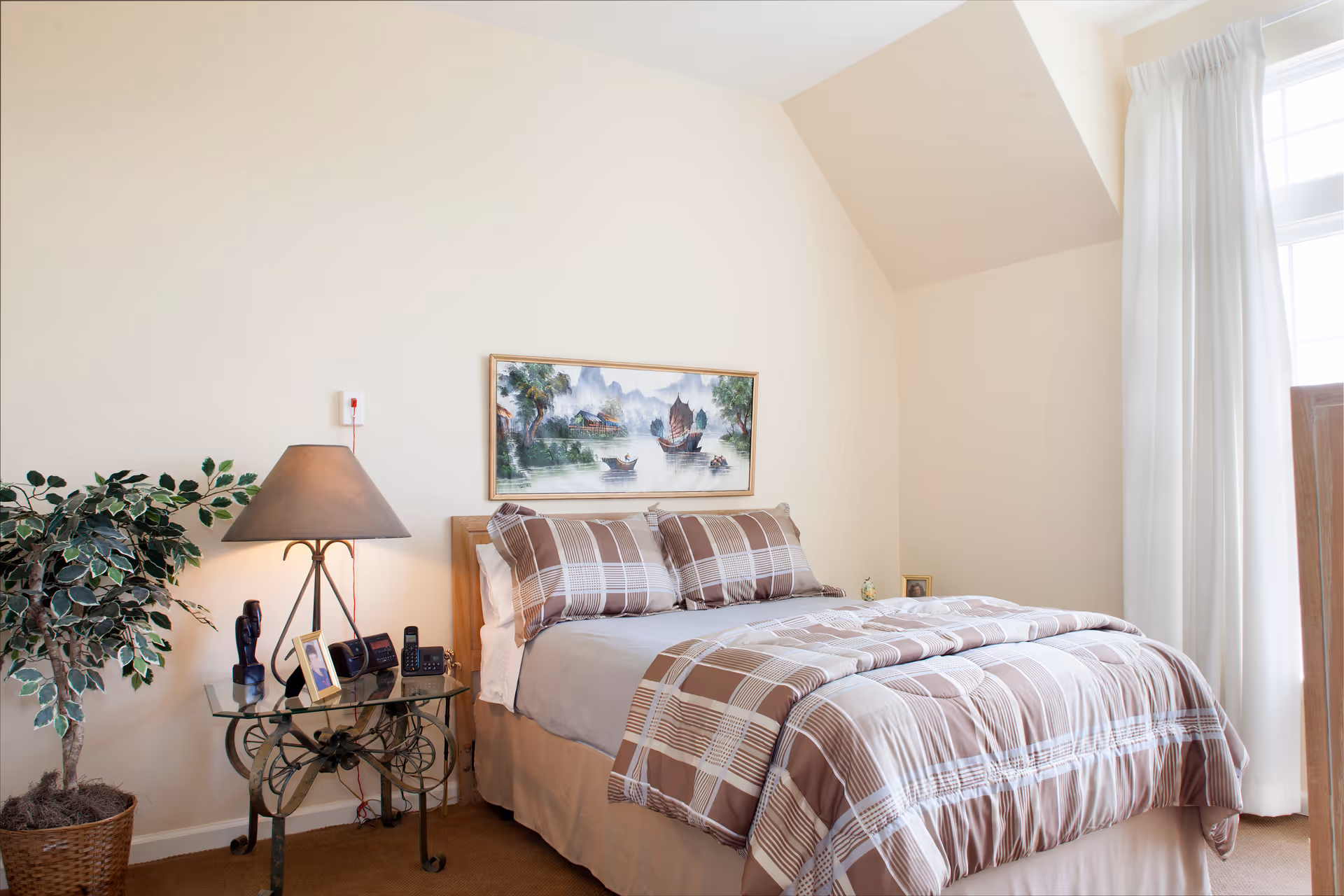 A neatly made bed with brown and beige plaid bedding in a bedroom. Next to the bed is a glass-top side table with a lamp, a potted plant, framed photos, and a telephone. A framed painting of boats on water hangs on the wall above the bed. White curtains cover a window on the right side of the room.