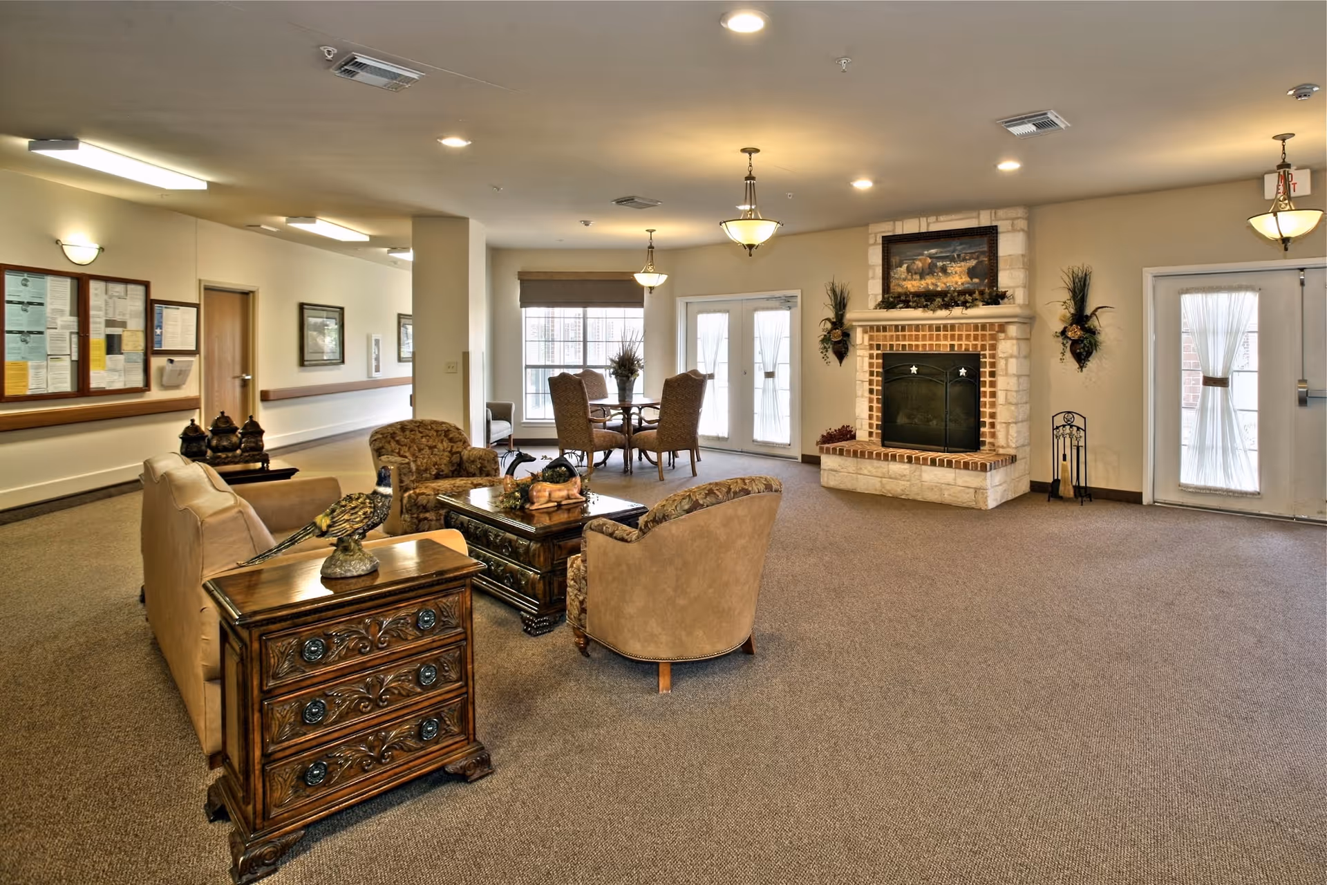 A spacious and warmly decorated living room area in a senior care facility featuring a stone fireplace with a painting above it, multiple upholstered chairs and sofas arranged around a wooden coffee table, a small dining table with four chairs near large windows, and soft lighting from ceiling fixtures.