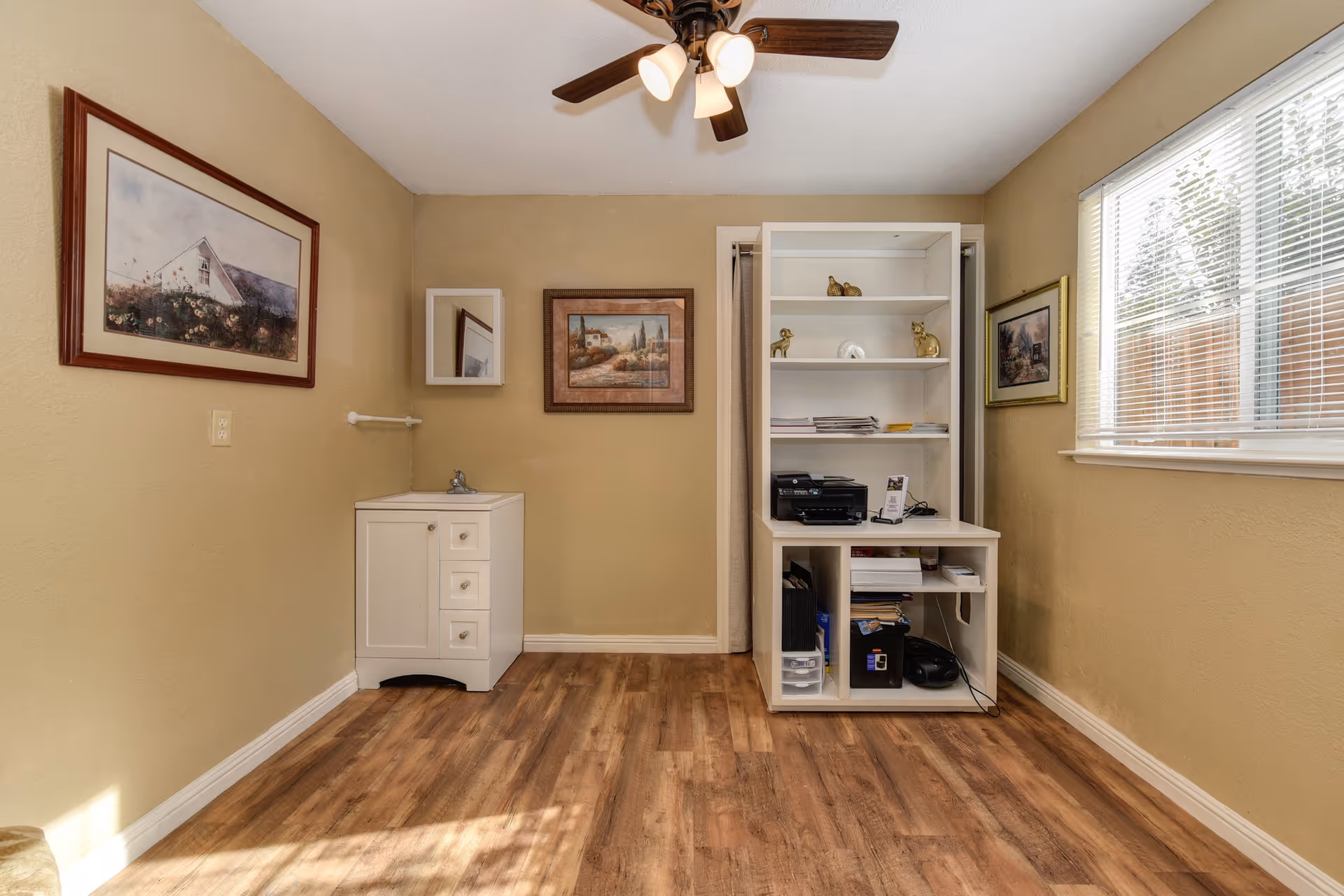 A small room with beige walls and wood flooring featuring a white cabinet with a sink on the left side, a white shelving unit with various items including a printer and decorative figurines on the right, three framed paintings on the walls, a window with blinds on the right, and a ceiling fan with lights above.