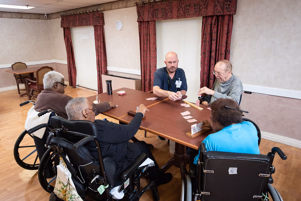 A group of elderly individuals in wheelchairs and a caregiver sitting around a wooden table playing a card game in a well-lit room with wooden flooring, beige walls, and red patterned curtains.