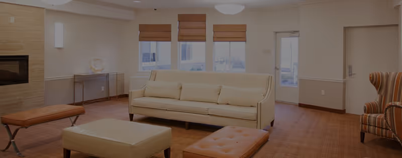 Sunlit living room with a beige sofa, ottomans, a striped armchair, and large windows.