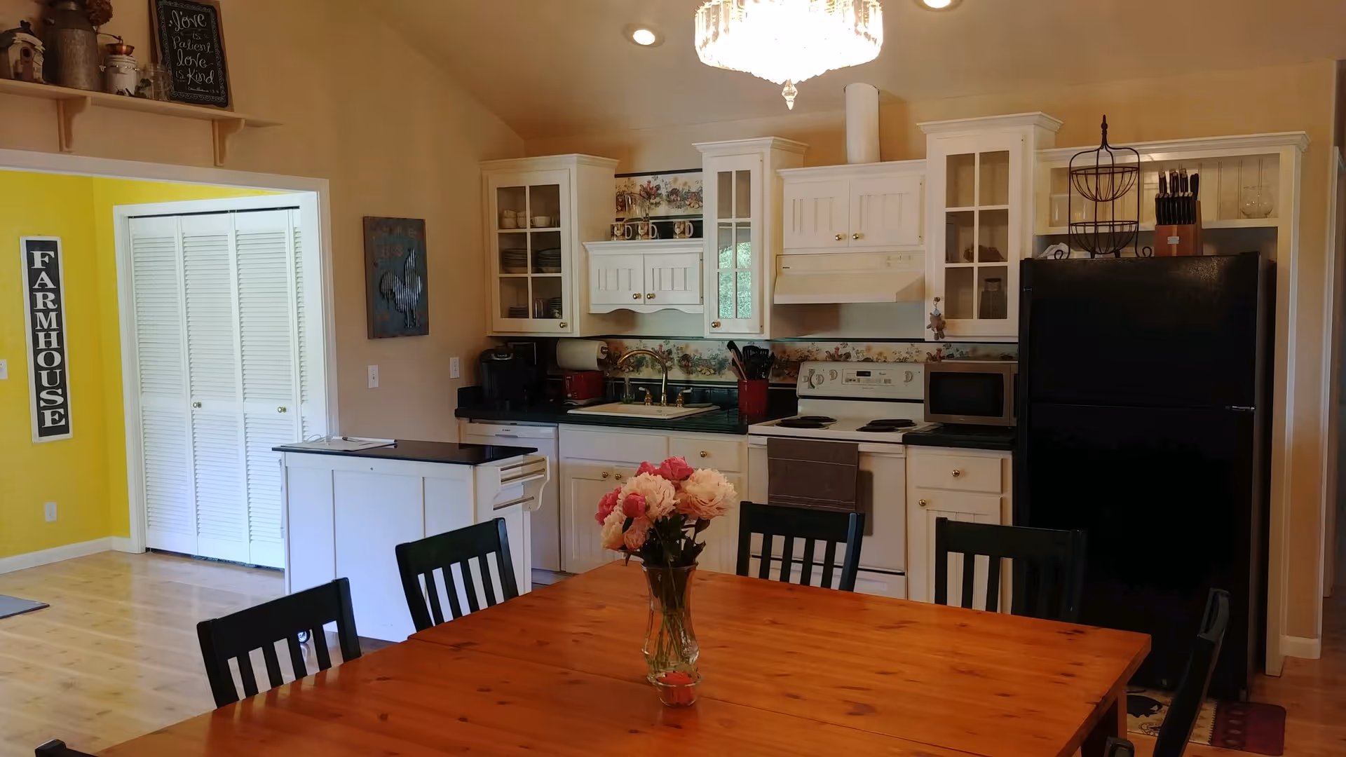 Open kitchen with white cabinets and a black refrigerator behind a wooden dining table topped with a vase of flowers.